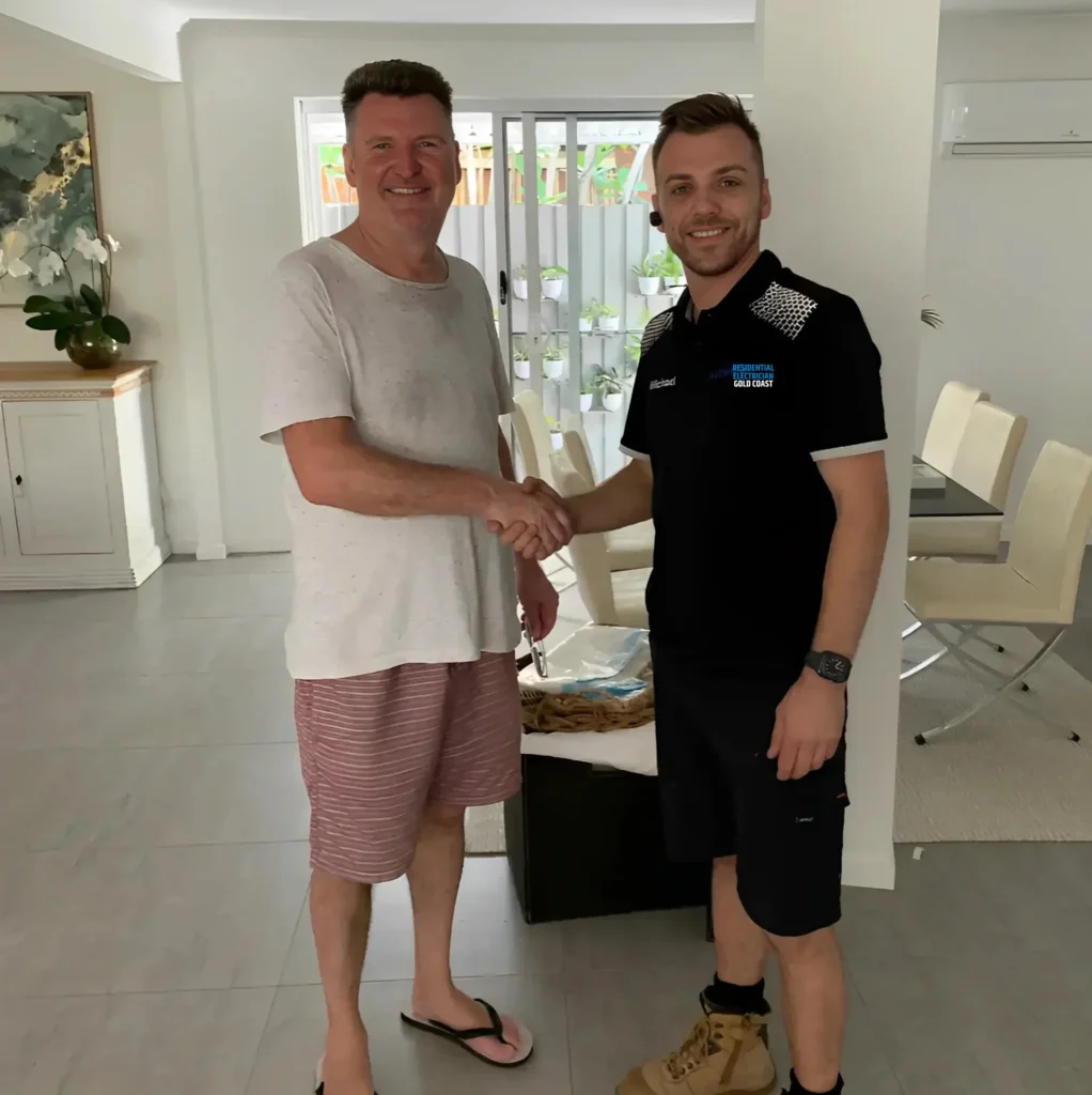Two people shaking hands inside a modern residential space. One wears a black uniform, the other a casual t-shirt and shorts.
